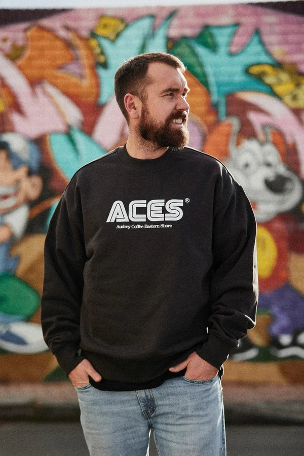 Man with hands in jean pockets, looking away from camera, wearing a black jumper with ACES Audrey Coffee Eastern Shore written on it