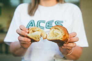 pastry being broken in half by a female. she's wearing an Audrey Coffee ACES white t-shirt